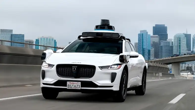 White self-driving car with roof sensors drives on a highway bridge with city skyscrapers in the background