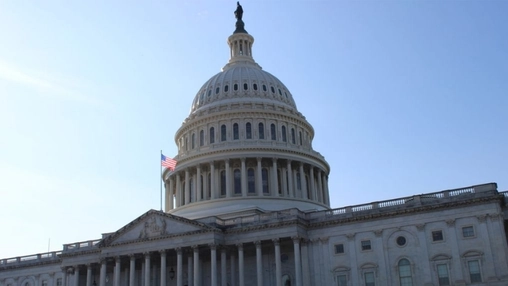 The dome and columned facade of the US Capitol building with an American flag flying against a clear blue sky